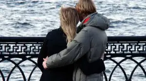Two people with light hair are standing by a decorative metal railing, embracing while looking out at a body of water. They are wearing jackets and standing close together, with their backs to the camera.