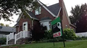 A brick house with white trim sits behind a For Sale sign marked SOLD on the front lawn, surrounded by greenery and trees. Steps lead to a small porch at the front entrance.
